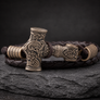 A Viking-style hammersmith bracelet featuring an intricately carved bronze-toned hammer head charm, knotted Celtic knot details, and textured metal end caps, resting on a dark brown braided leather cord over a dark slate surface.