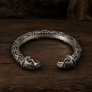 An ornate silver-toned open cuff bracelet with detailed Norse-style animal head motifs at both ends, displayed on a dark rustic wooden surface.