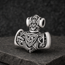 An antiqued silver Viking-style mace pendant with intricate Celtic knotwork and a triquetra symbol, displayed on a dark textured stone surface.