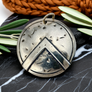 Silver pendant with Spartan shield with geometric design on a dark surface with green leaves