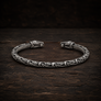 An oxidized sterling silver Viking-style cuff bracelet with intricate knotwork and wolf head closures, displayed on a dark rustic wooden surface.