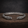 An oxidized silver Viking-style cuff bracelet with detailed carvings and fish-head motifs on both ends, displayed on a dark wooden surface against a dark textured background.