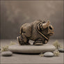 A small textured metal bull figurine with an antique bronze finish, resting on a smooth gray stone surrounded by decorative moss and stones against a soft gray background.