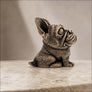 A small sculpted dog figurine with a textured metallic finish, sitting on a light stone surface against a soft neutral background.