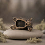 An antique bronze-finished metal bear figurine lying on its back, resting on a smooth gray stone with blurred dried flowers in the background.