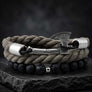 A braided rope bracelet with a Viking-style axe clasp, resting on a dark slate surface. The top layer is a thick taupe-brown braided cord, and the bottom layer consists of small black lava beads with a silver spacer.