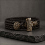 A dark brown braided leather wrap bracelet with an ornate bronze-toned metal clasp shaped like a Viking hammer, displayed on a dark stone slab against a textured gray background.