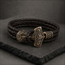 A dark brown braided leather bracelet featuring a detailed bronze Viking-style hammer clasp, displayed on a dark slate surface.