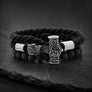 A black braided cord bracelet with dark porous beads and a central engraved silver-toned cross clasp, displayed on a dark stone surface.