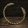 A twisted metal cuff bracelet with Viking raven bird head ends, featuring a textured bronze and gold finish, displayed on a rustic dark wood background.