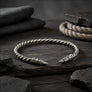 A sterling silver twisted bracelet with ornate engraved raven head ends, displayed on a dark wooden surface with dark fabric and stones in the background.