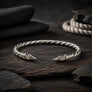 A sterling silver twisted bracelet with ornate engraved raven head ends, displayed on a dark wooden surface with dark fabric and stones in the background.
