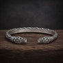 An oxidized silver Viking-style cuff bracelet with detailed carvings and fish-head motifs on both ends, displayed on a dark wooden surface against a dark textured background.