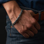 A close-up of a man's hand and wrist wearing a silver braided metal cuff bracelet, paired with blue jeans and a brown leather belt.