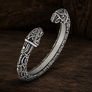 An ornate silver-toned open cuff bracelet with detailed Norse-style knotwork engravings and animal head motifs on both ends, displayed on a dark wooden surface.