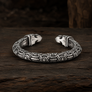 An ornate silver-toned cuff bracelet with intricate engraved patterns and raven head closures, displayed on a dark rustic wooden surface.