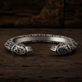 A silver-toned open cuff bracelet with intricate Norse-style knotwork and raven carvings on both ends, displayed on a dark rustic wooden surface.