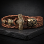 A Norse-style braided leather bracelet with an engraved bronze-toned hammer-shaped clasp and matching decorative beads, displayed on a dark stone slab against a dark background.
