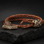A braided brown leather bracelet with an antique bronze-toned hammer-shaped charm and matching decorative bead, displayed on a dark stone surface against a dark background.