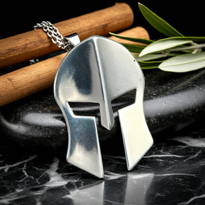 Silver Spartan helmet pendant on a dark surface with wooden sticks and leaves.