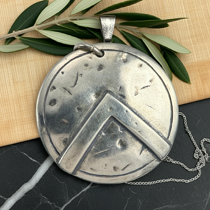 Silver Spartan shild pendant with geometric design on a dark surface with green leaves