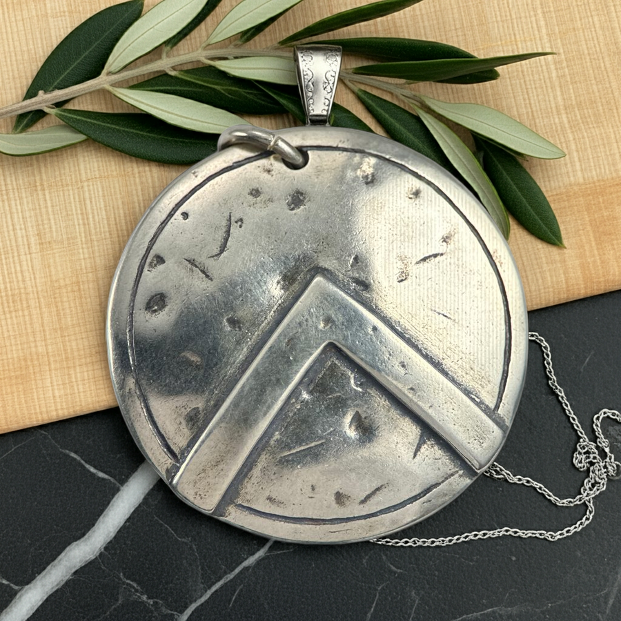 Silver Spartan shild pendant with geometric design on a dark surface with green leaves