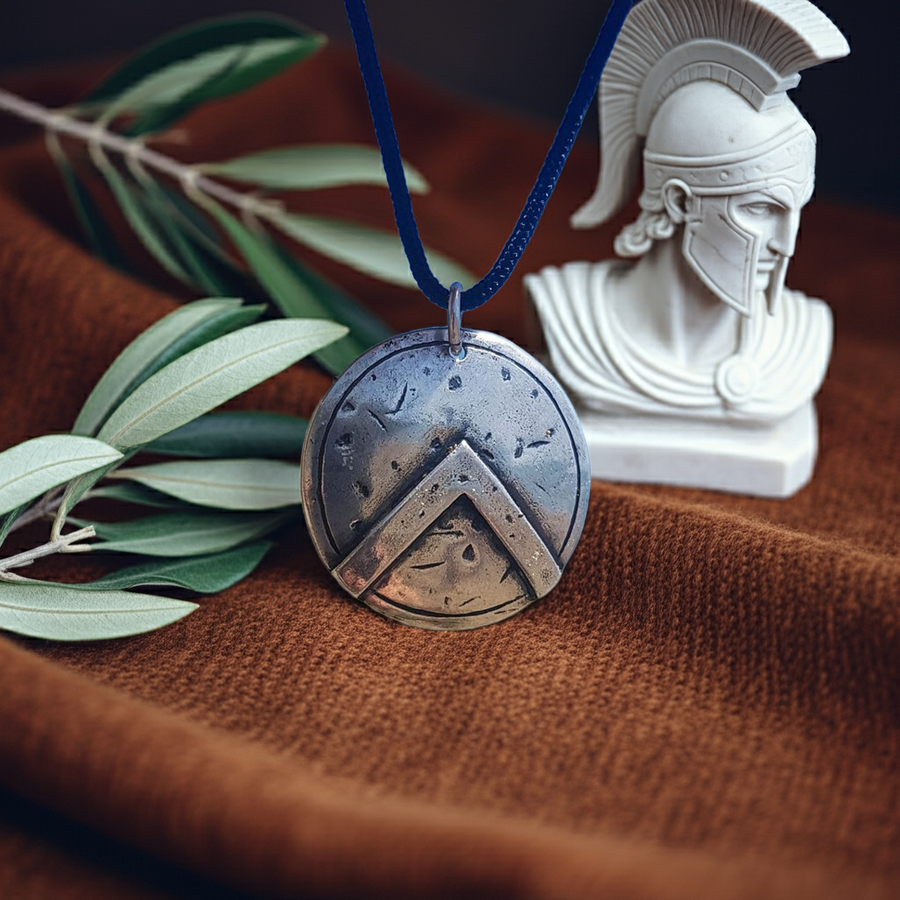Silver Spartan shield pendant on a blue cord with a white warrior statue and olive branch on a brown surface