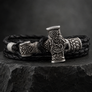 A black braided leather bracelet with an intricately carved silver-toned metal hammer-shaped clasp, displayed on a dark textured stone surface.