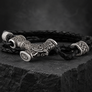A black braided leather bracelet with an ornate silver-toned metal centerpiece featuring Celtic or Norse-style knotwork and a Viking horn-like charm, displayed on a dark textured stone surface.