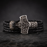 A black braided leather wrap bracelet with an ornate silver-toned Celtic knot and Viking-style hammer-shaped clasp, displayed on a dark textured stone surface.