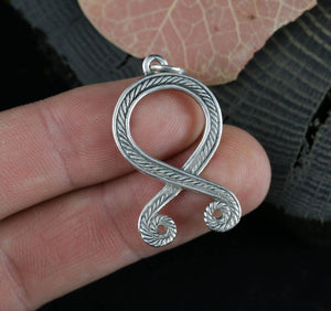 Silver Troll Cross pendant held between fingers with a leafy background