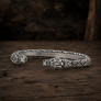 A silver-toned open cuff bracelet with carved tribal animal head motifs on both ends and a braided pattern along the band, displayed on a rustic dark wood surface.