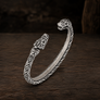 An open cuff bracelet made of oxidized silver with detailed Norse-style knotwork and animal head motifs at both ends, displayed on a dark wooden surface.