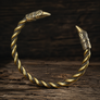 A twisted metal cuff bracelet with Viking raven bird head ends, featuring a textured bronze and gold finish, displayed on a rustic dark wood background.