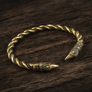 A twisted metal cuff bracelet with viking raven ends, shown on a dark wooden surface.
