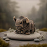 A small metallic bull figurine with a textured bronze finish, resting on a smooth round stone pedestal. The scene is styled with moss, small stones, and delicate dried flowers against a soft, blurred forest background.