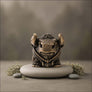 Bronze-finished bull figurine sitting on a round gray stone pedestal, surrounded by small stones and dried greenery against a soft blurred gray-brown background.