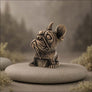 A small bronze-finished French bulldog figurine sits on a smooth round stone in a softly blurred outdoor-style setting with muted greenery and trees in the background.