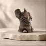 A small sculpted dog figurine shown from the back, sitting on a light stone slab against a soft beige studio background.