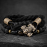 A black braided cord bracelet with matte black porous beads, a central Viking-style hammer charm in antiqued gold-tone metal, and two gold-tone cylindrical end caps, displayed on a dark slate surface.