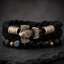 A black braided cord bracelet with ornate bronze Viking-style knotwork charms, cylindrical bronze beads, and black stone beads, displayed on a dark slate surface.