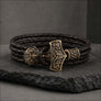 A dark brown braided leather wrap bracelet with an ornate bronze-toned Viking-style hammer clasp, displayed on a dark gray stone slab.