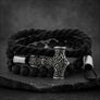 A black braided cord bracelet with a silver-toned engraved hammer charm, black beads, and silver end caps, displayed on a dark stone surface.
