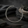 A sterling silver twisted bracelet with ornate engraved raven head ends, displayed on a dark wooden surface with dark fabric and stones in the background.