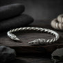 A sterling silver twisted bracelet with ornate engraved raven head ends, displayed on a dark wooden surface with dark fabric and stones in the background.