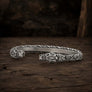 A silver-toned open cuff bracelet with carved tribal animal head motifs on both ends and a braided pattern along the band, displayed on a rustic dark wood surface.