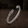 An open cuff bracelet made of oxidized silver with detailed Norse-style knotwork and animal head motifs at both ends, displayed on a dark wooden surface.